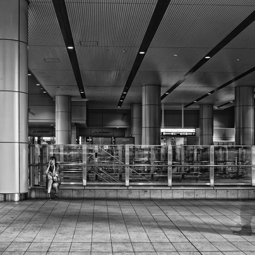 A man, his back against a pillar, looking to his right. A couple walk from right to left, they are blurry due of the shutter speed. Black and white picture.
